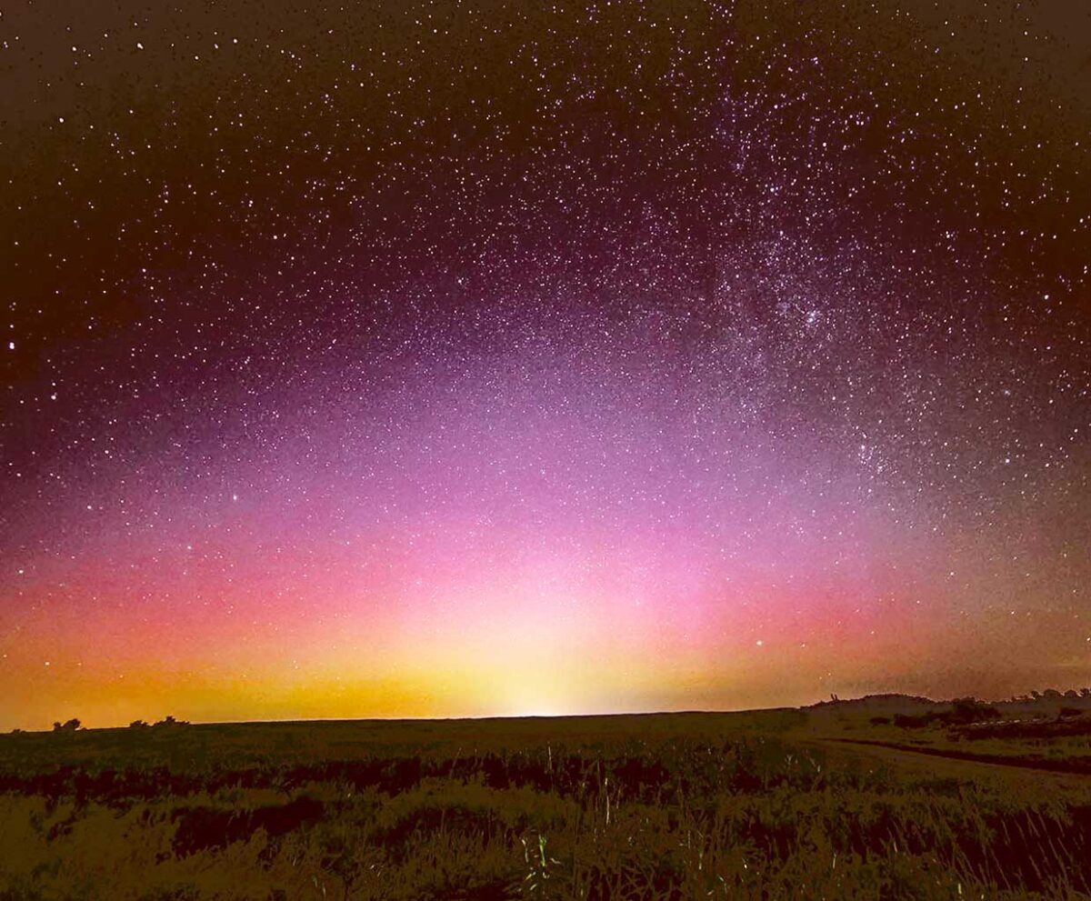 Northern lights glow softly over a vast horizon, fading from golden yellow to pink and purple under a star-speckled sky